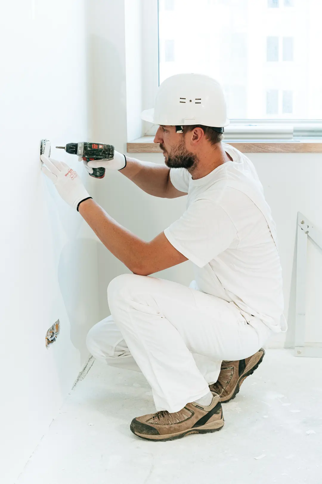 Artisan casqué et ganté, accroupi, fixant un appareillage électrique mural à la visseuse dans une pièce repeinte