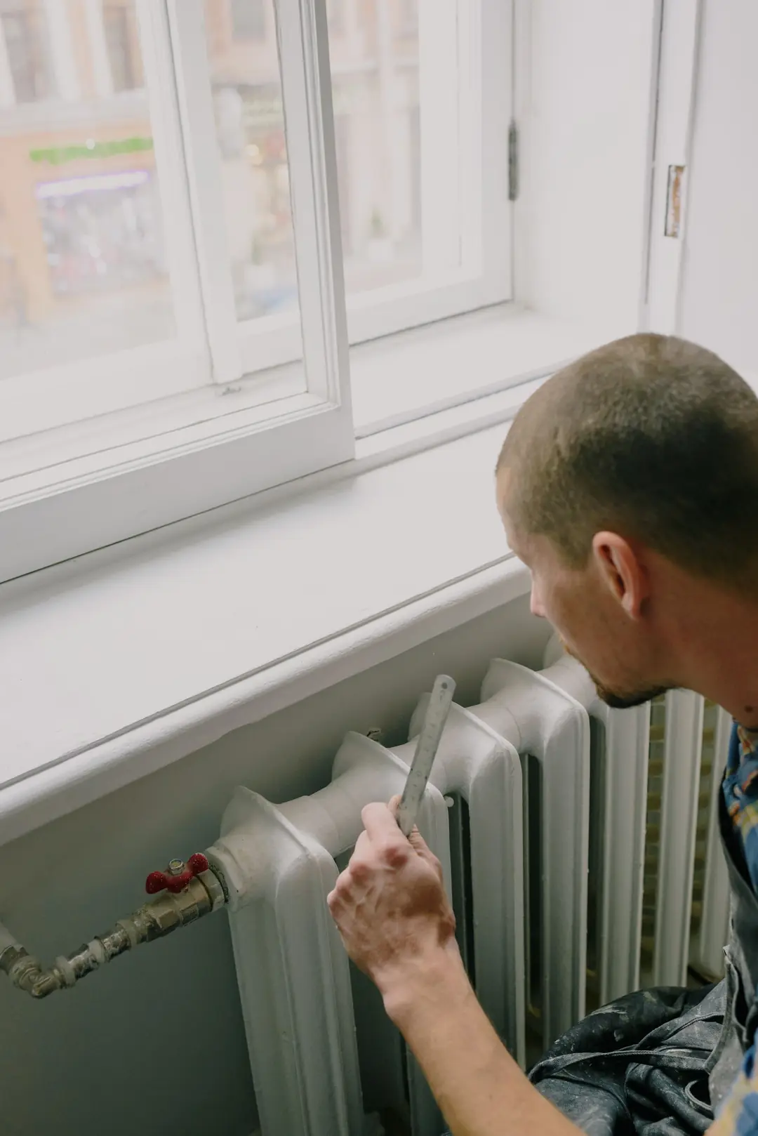 Artisan mesurant à la règle un radiateur en fonte sous une fenêtre, à côté de sa vanne d’arrivée